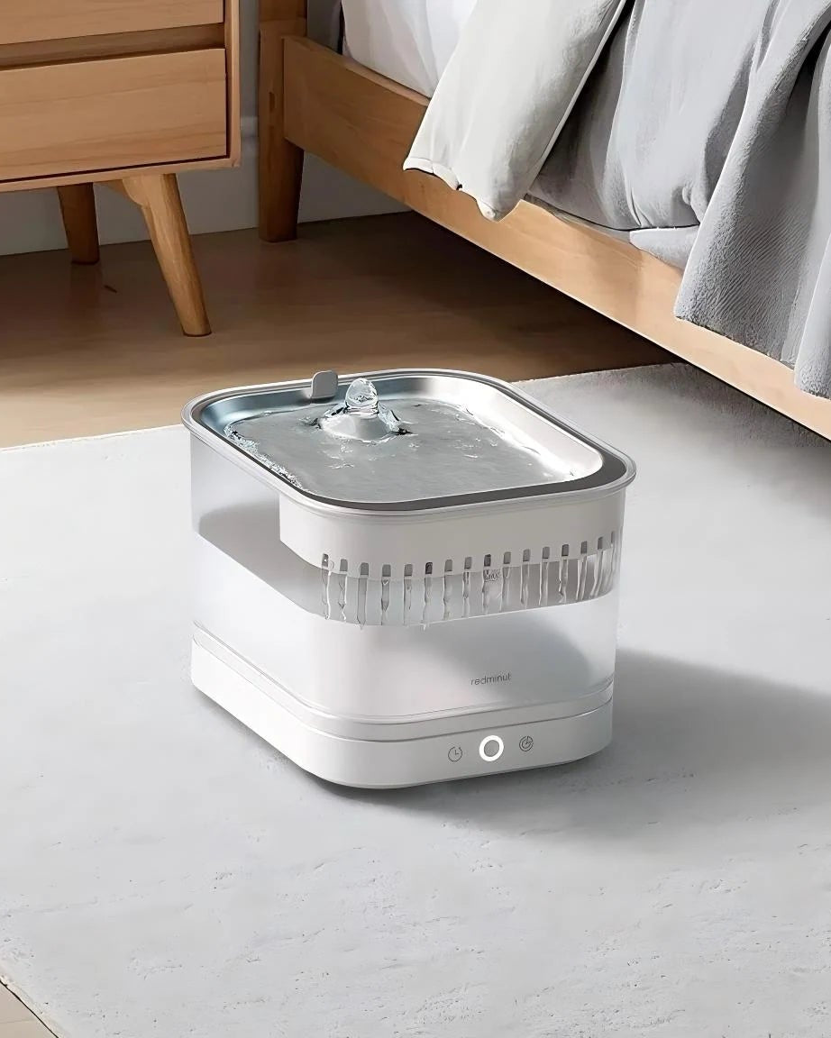 White pet water fountain on a light-colored floor with a wooden bed and side table in the background.